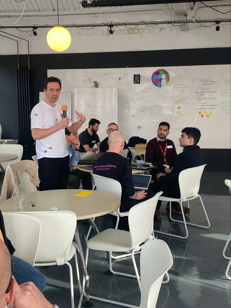 Participant talking into a microphone with other participants sat at a table. White boards in the backgroun