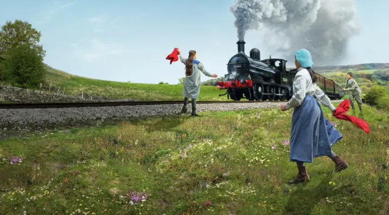 Three children, in early 20th century clothing, flag down a steam train in the Yorkshire countryside.